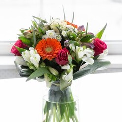 In this stunning arrangement from a Greenwich florist, the artistry of colour and structure meets a sense of local charm. The bouquet is anchored by creamy-beige gerbera daisies, their delicate petals fanning out in gentle halos around brilliant orange counterparts-vivid reminders of the glowing skies over Greenwich at dusk. Plush, fuchsia roses are interwoven, their bold colour bringing a sense of depth and passion that speaks to Greenwich's dynamic community spirit. Interspersed throughout are white alstroemeria and dainty spray roses, providing crisp contrast and a sense of lightness, while traces of soft pink blossoms infuse subtle warmth. The substantial presence of broad, dark green aspidistra leaves, precisely folded to form a protective collar, grounds the effervescent blooms in lush tranquility-much like the sheltering trees of Greenwich Park. Surrounding these, elegant pointed leaves and wispy sprigs of greenery complete the composition, introducing further depth and natural movement. Housed in a sleek, clear glass vase, the bouquet's vibrant green stems are visible beneath the water line, enhancing its sense of fresh vitality. Soft daylight seems to brighten the bouquet without overpowering it, casting each petal in a glow that recalls the peaceful mornings along the Thames. This is a bouquet made for celebration-a fitting centrepiece for gatherings in Greenwich, a beautiful way to mark a milestone, or a heartfelt gesture for new adventures.
